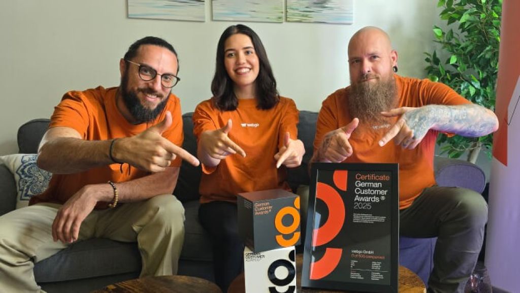Drei Mitarbeiter der webgo GmbH sitzen auf einer Couch. Sie haben alle orangenen Shirts an, worauf das webgo Logo in weiß zu sehen ist. Sie freuen sich über den Erhalt des German Customer Award und zeigen alle auf die German Customer Award-Skulptur sowie ein eingerahmten Zertifikat, welche vor Ihnen auf einem Tisch abgelegt sind.