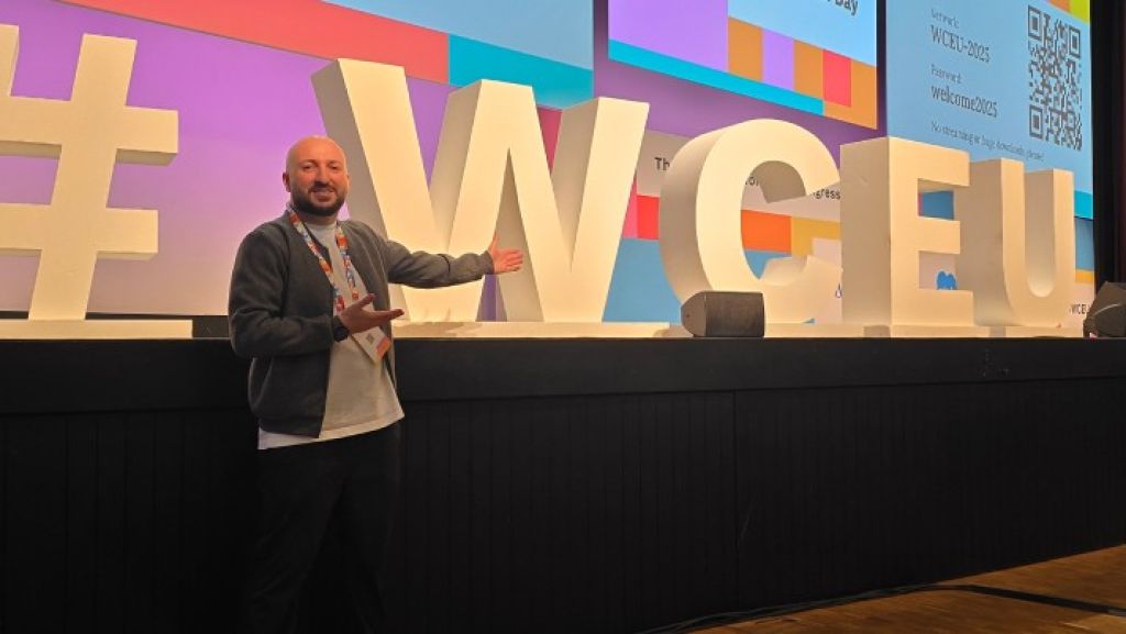 Serhat Köylüce, Online Marketing Manager der webgo GmbH, während des Contribution Day auf dem WordCamp Europe 2025 in Basel. Er zeigt auf eine Schrift-Skulptur, die auf einer Bühne steht und worauf #WCEU steht.