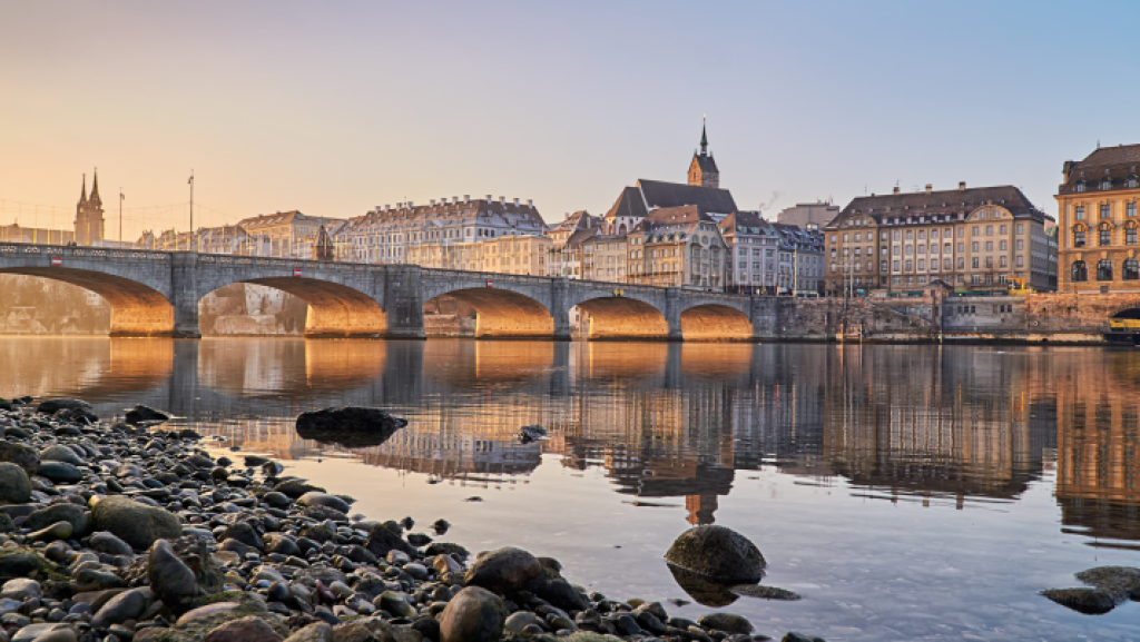 Blick auf die Mittlere Brücke und die Altstadt von Basel bei Sonnenuntergang mit ruhigem Rhein im Vordergrund.