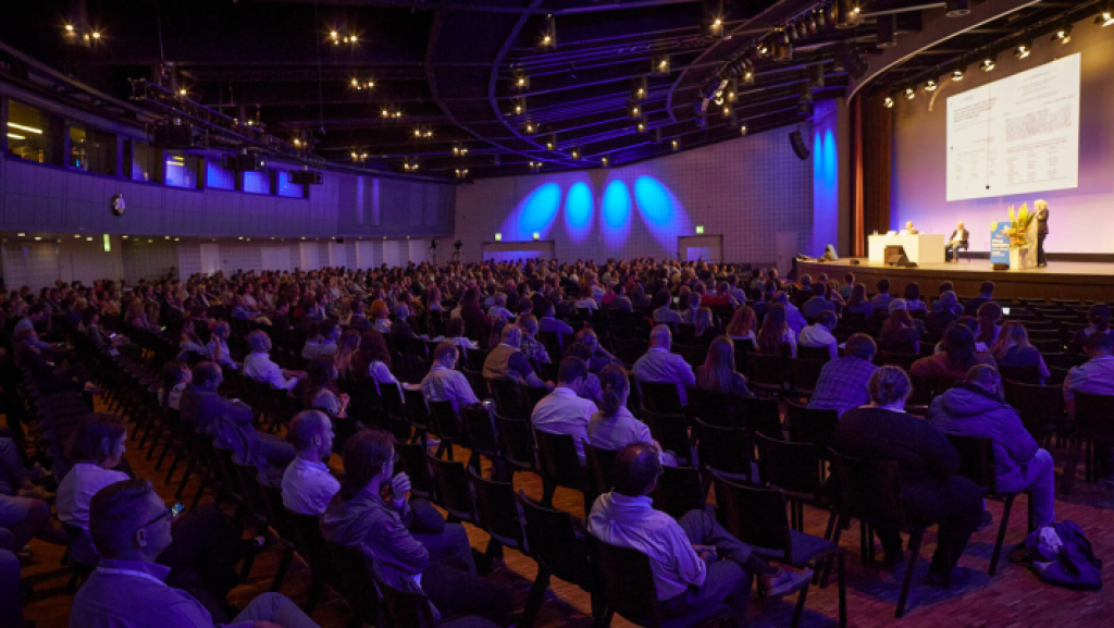 Großes Auditorium in der Messe Basel: Zahlreiche Besucherinnen und Besucher verfolgen konzentriert eine Präsentation. Die Bühne ist in Blau- und Violetttönen ausgeleuchtet.