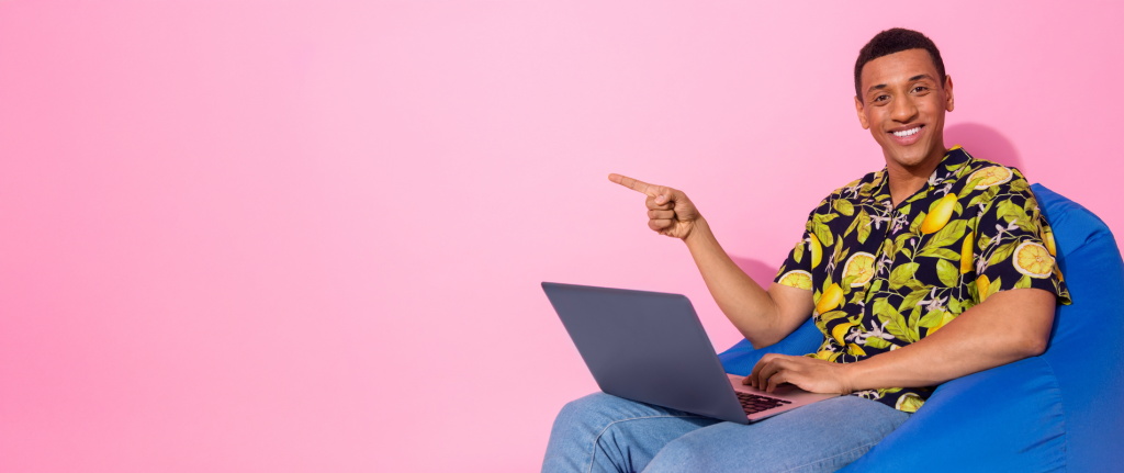 Junger Mann sitzt entspannt auf einem blauen Sitzsack vor rosa Hintergrund, arbeitet mit einem Laptop und zeigt lächelnd zur linken Bildhälfte – symbolisiert unkompliziertes, leistungsstarkes NVMe Webhosting von webgo mit hoher Benutzerfreundlichkeit.