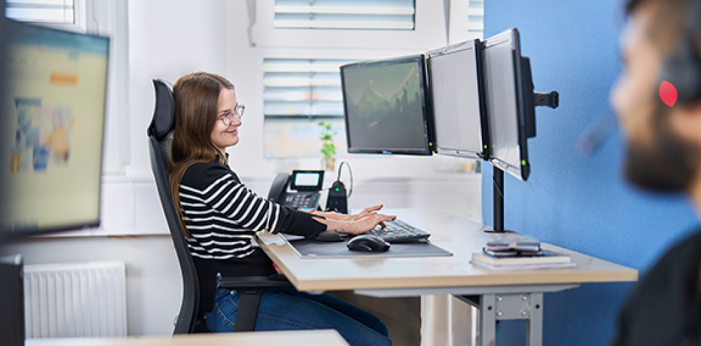 In den Büro-Räumlichkeiten der webgo GmbH ist eine Kundensupport-Mitarbeiterin, welche an Ihrem Arbeitscomputer ein Kundenanliegen bearbeitet. Ein zweiter Mitarbeiter ist ebenfalls zu sehen, welcher ebenfalls an seinem Arbeitscomputer sitzt und mit einem Kunden telefoniert, um diesem bestmöglich zu unterstützen.